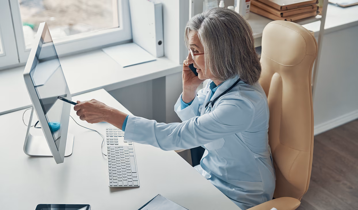 Senior female doctor with gray hair wearing a white coat and stethoscope, sitting at a desk, talking on the phone and pointing at a computer monitor in a bright office.