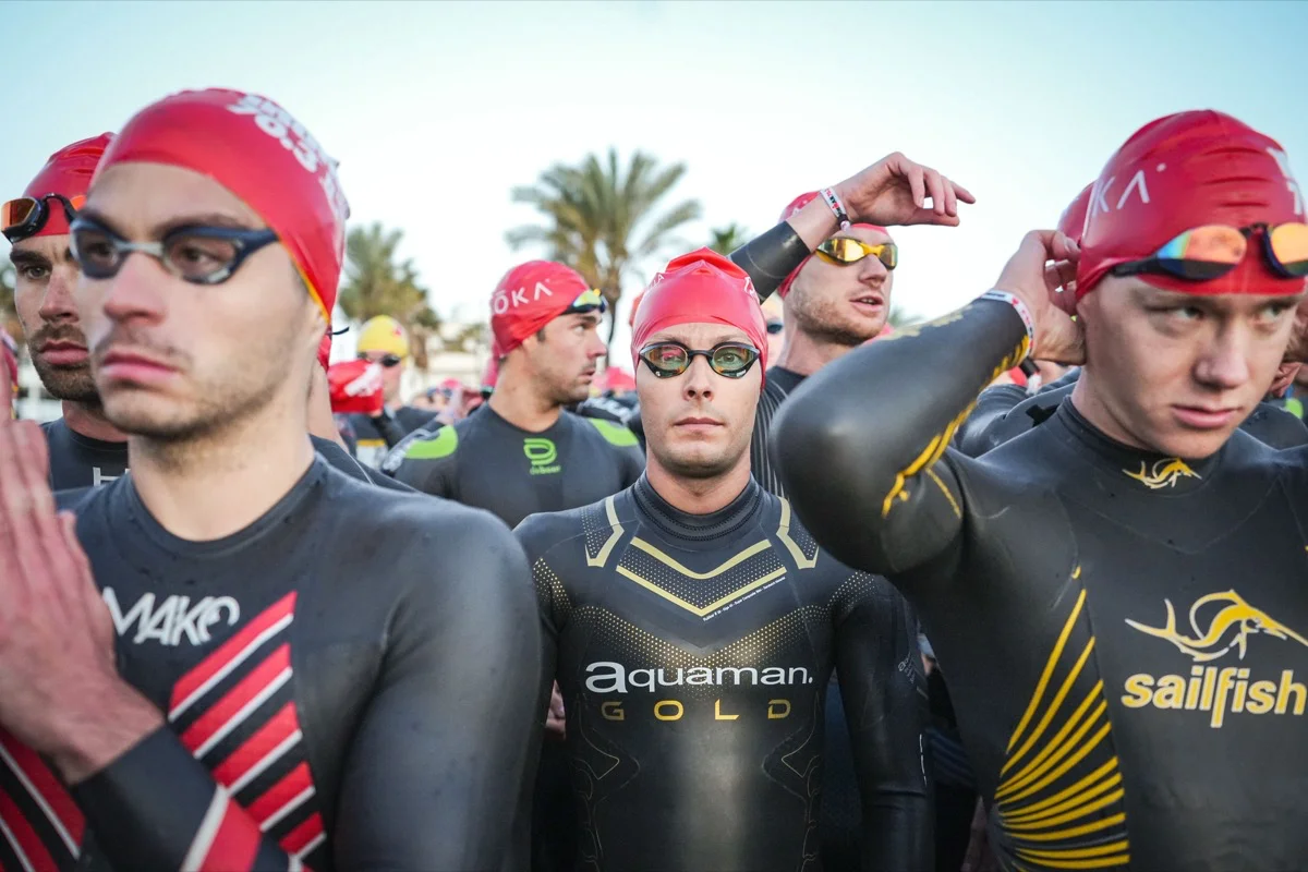 Athleten in Neoprenanzügen und roten Badekappen bereiten sich vor einem Wettkampf auf das Schwimmen vor.