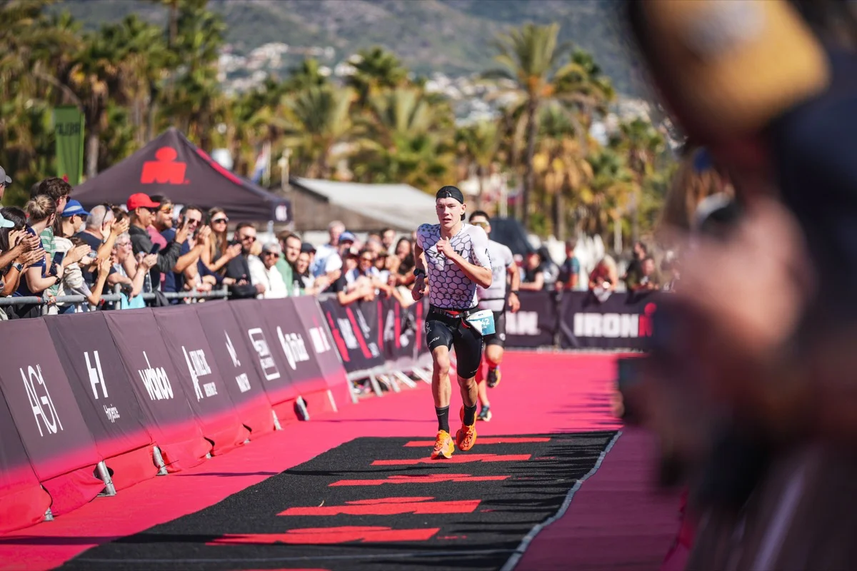 Athlet beim Laufen auf einer roten Strecke beim Ironman-Wettbewerb, umgeben von jubelnden Zuschauern und Palmen im Hintergrund.