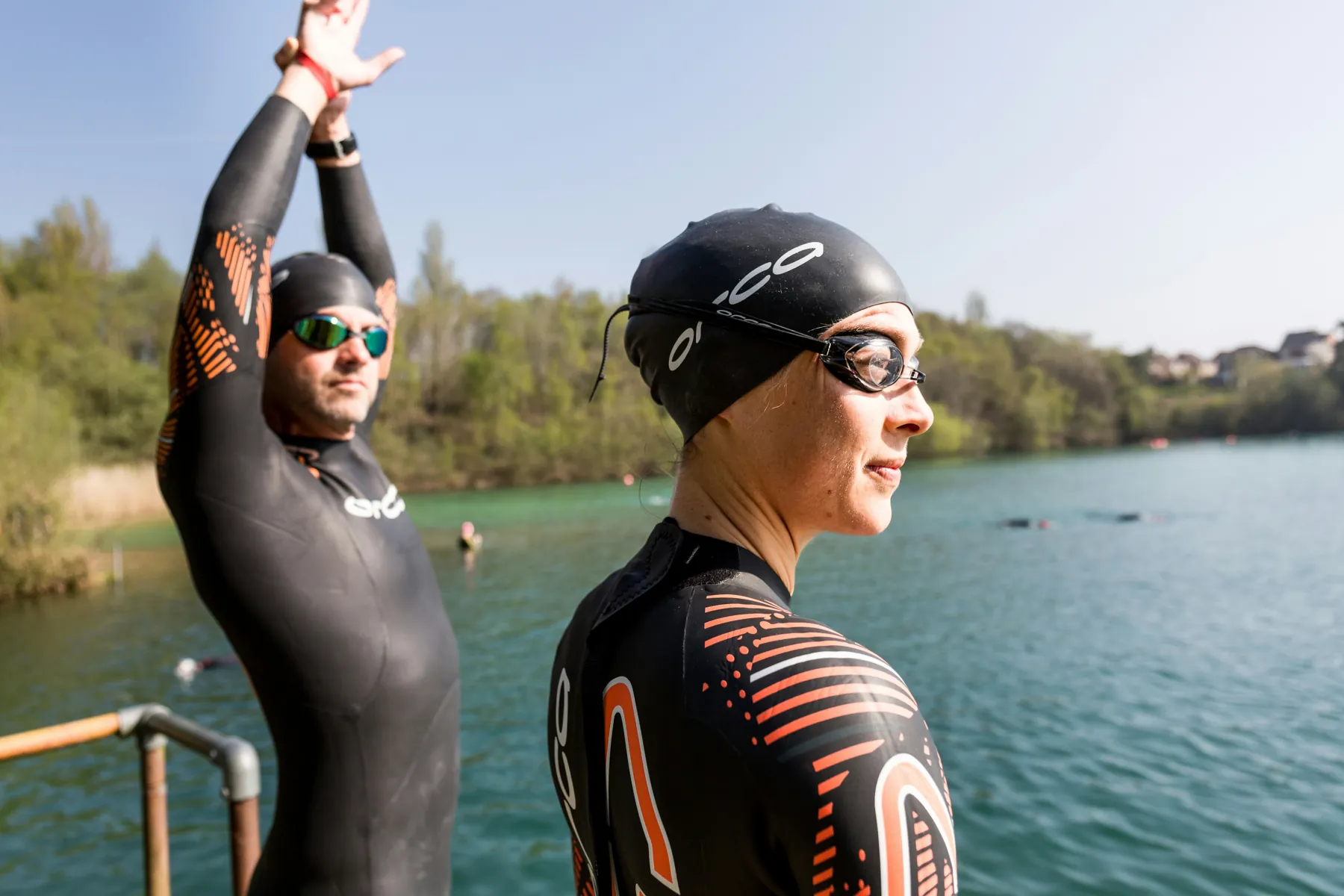 Zwei Triathlet:innen im Neoprenanzug warten am Steg eines Sees auf den Schwimmstart