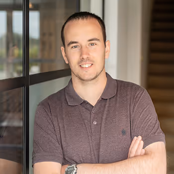 Smiling person in brown polo shirt standing near glass window