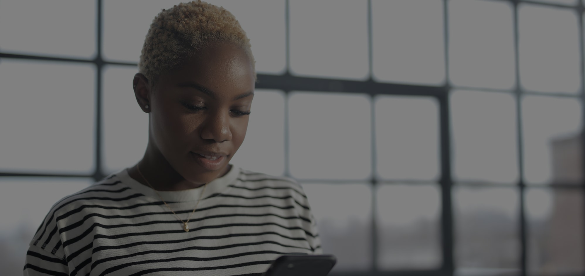 Person with short hair in striped shirt looking at phone in front of large windows