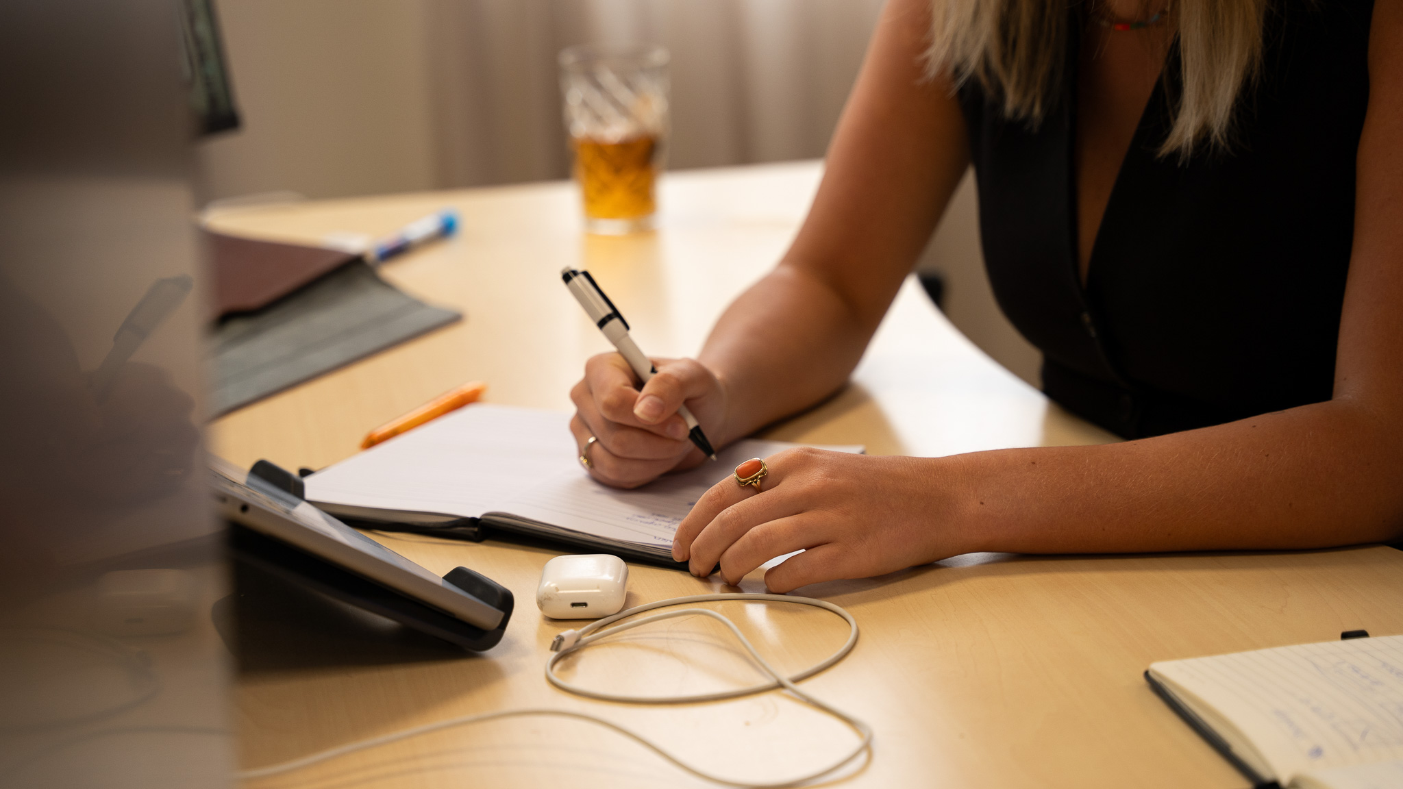 Vrouw schrijft notities in schrift op bureau met glas en oordopjes erbij.
