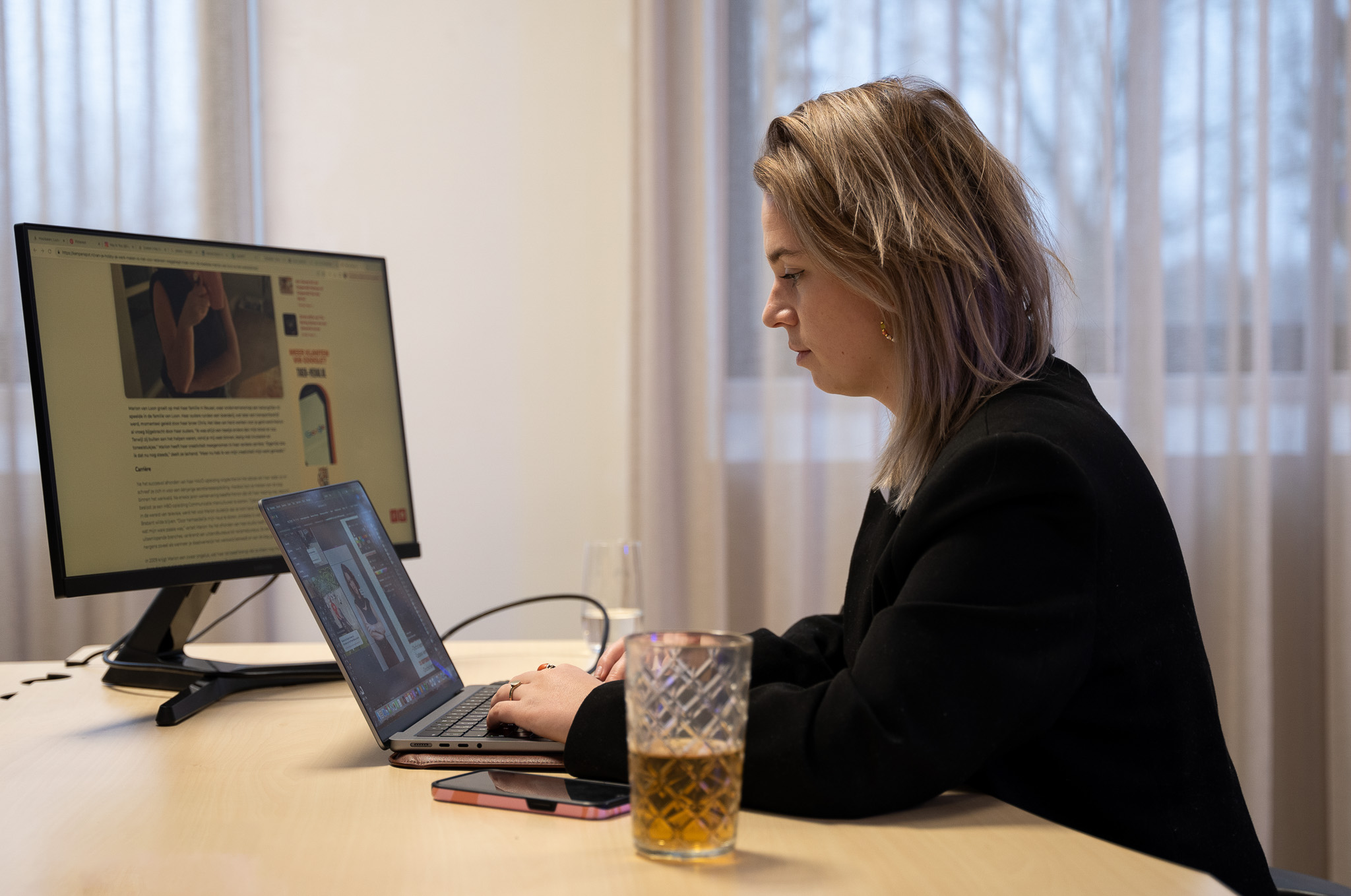 Vrouw werkt geconcentreerd op laptop aan bureau met drankje erbij.