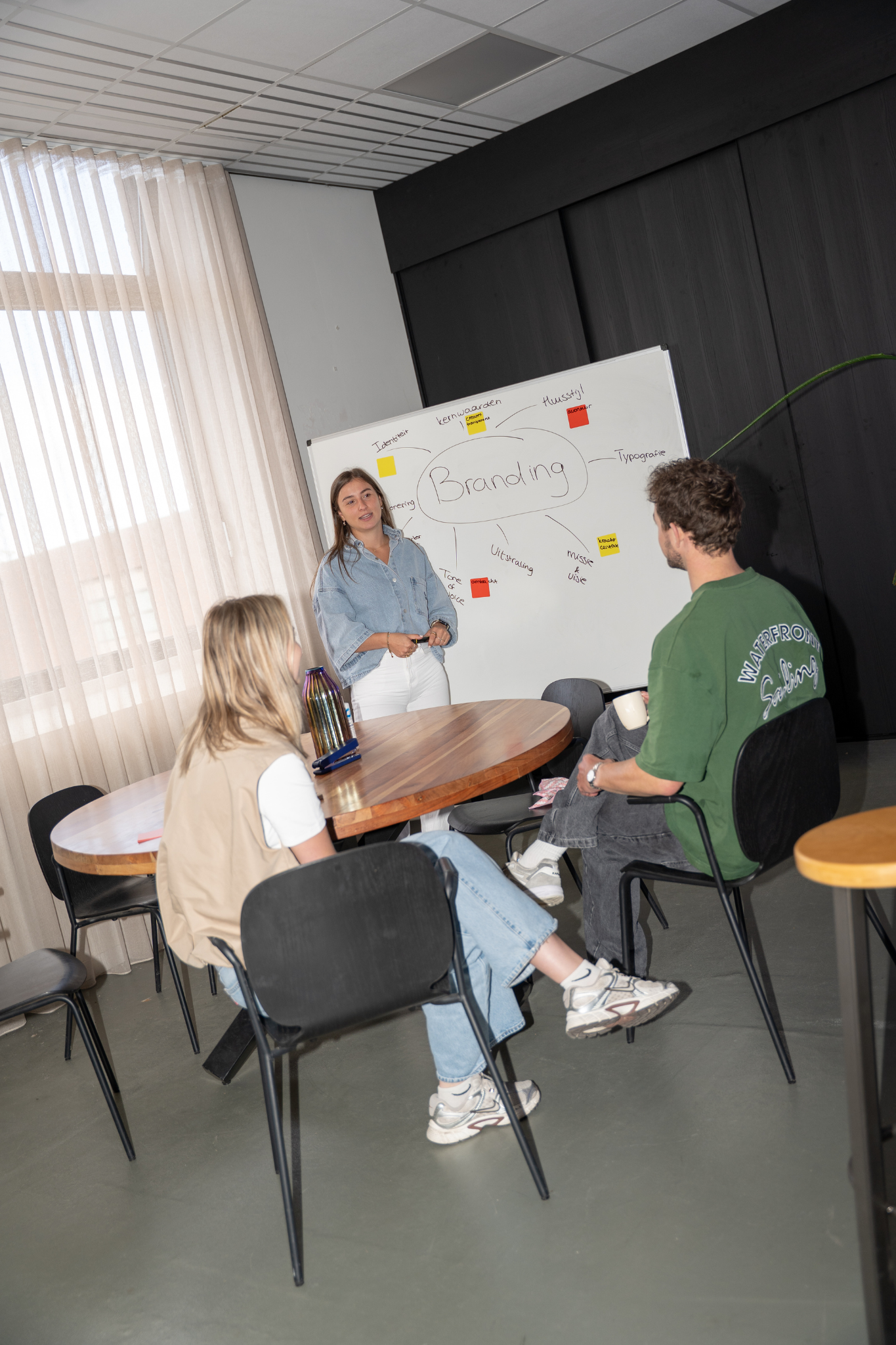 Vrouw geeft presentatie over branding aan twee collega’s bij een whiteboard.