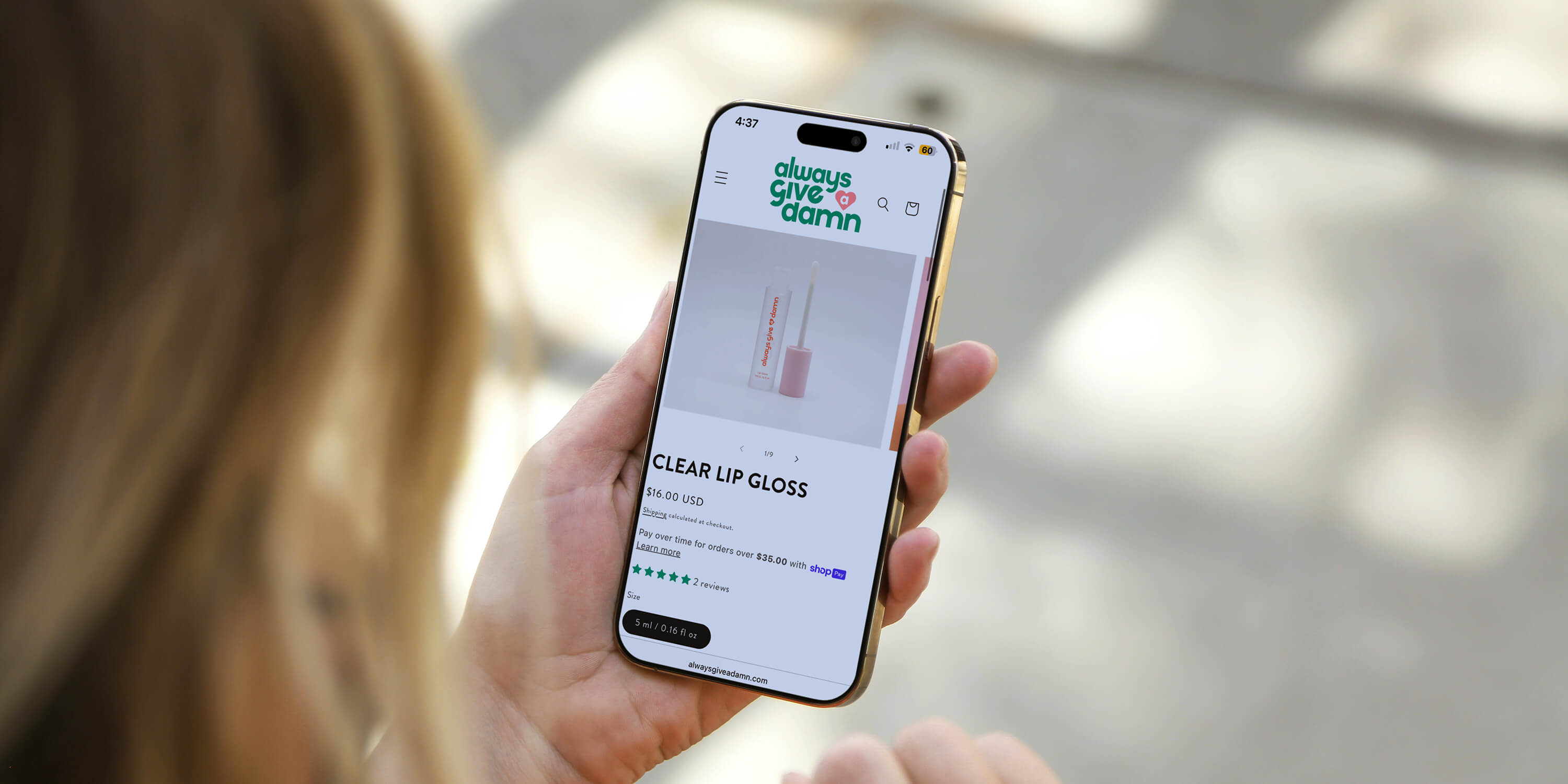 Mobile website mockup of woman holding a phone showing the Always Give A Damn product page on the website for the clear lipgloss designed by AP The Creative. 
