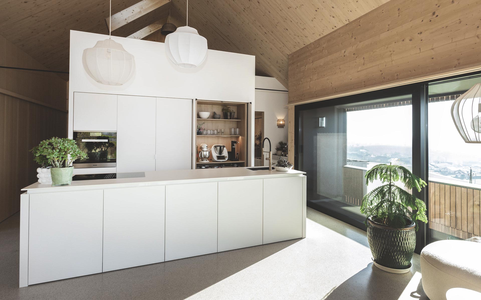 Eine moderne Holzküche mit weißen Holzplatten verkleidet befindet sich in einem großen mit natürlichem Holz ausgekleideten Wohnraum. Die großen Fenster lassen sehr viel Licht herein, was den Raum sehr freundlich erscheinen lässt. 
