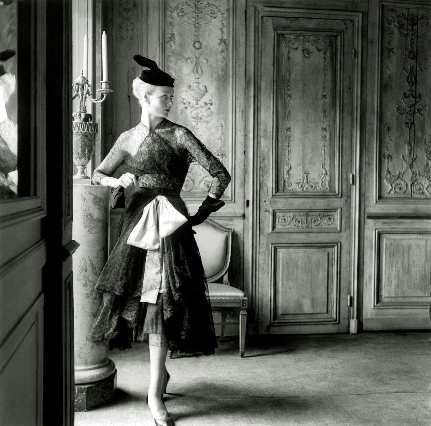 A model in a dress with a lacy, somewhat mesh bodice and layered, lacy skirt with a large bow dangling from the waist, heels, a hat, gloves (one on and the other in her hand) inside an ornate room.
