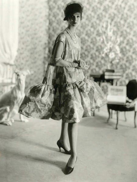 A model in a floral dropped waist baby doll dress, gloves, heals and a hat in a decorated room.
