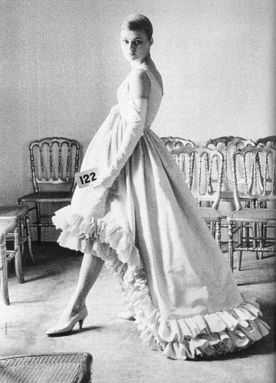 A model in a peacock dress with ruffes along the hem, gloves and heels holding a card with 122 written on it in a room in front of some chairs.