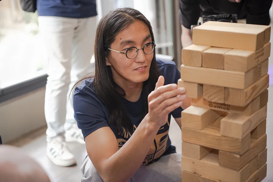 Young person with long hair and round glasses carefully pulling a wooden block from a large Jenga tower during a game indoors.