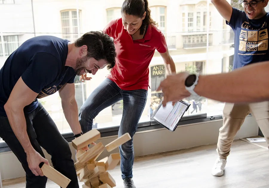 Three people indoors enthusiastically playing a game with wooden blocks, with one man bending down as blocks fall, a woman in a red shirt smiling, and another person cheering with arms raised.