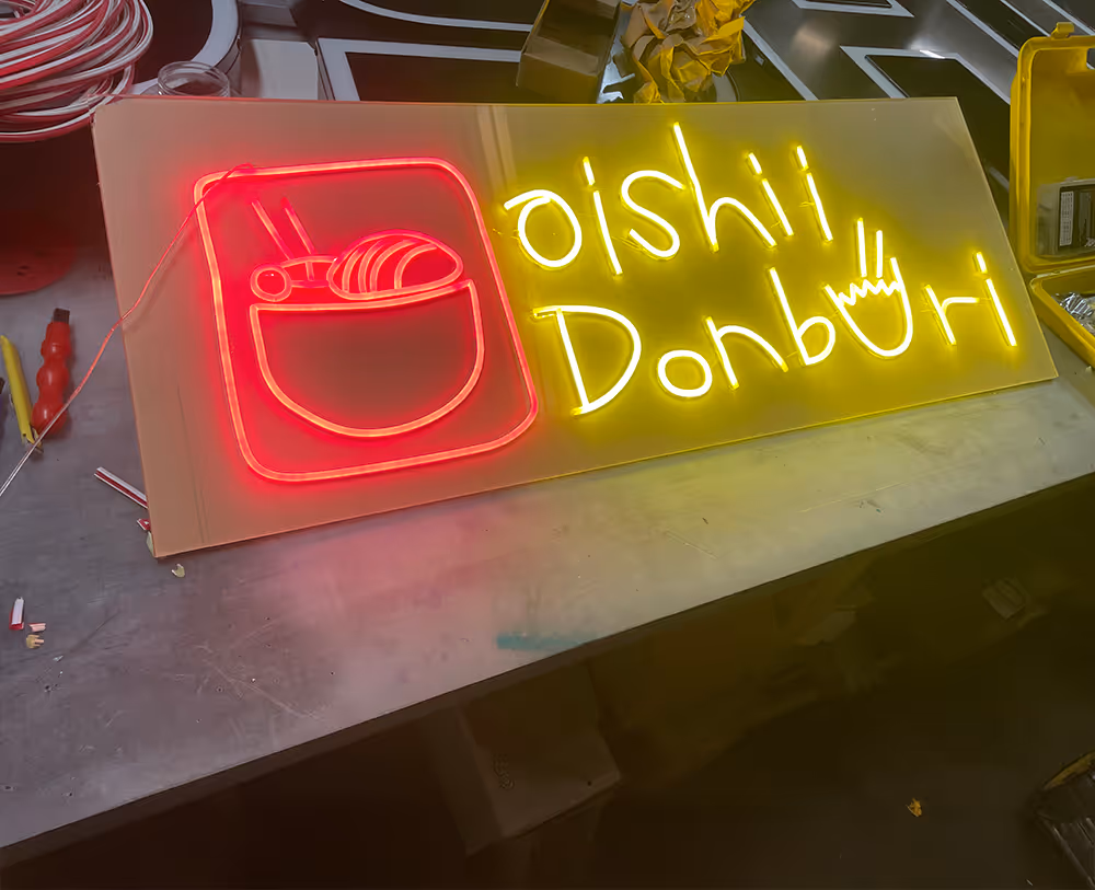 Neon sign on workbench displaying a red bowl with chopsticks and sushi, and yellow text reading 'oishii Donburi' with a bowl icon replacing the letter 'u'.