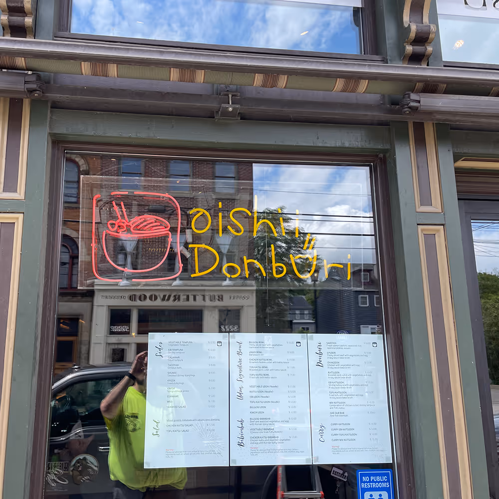 Shop window of Oishii Donburi featuring a neon sign with a bowl and chopsticks, three menus displayed, and a reflection of a building and person in a yellow shirt.