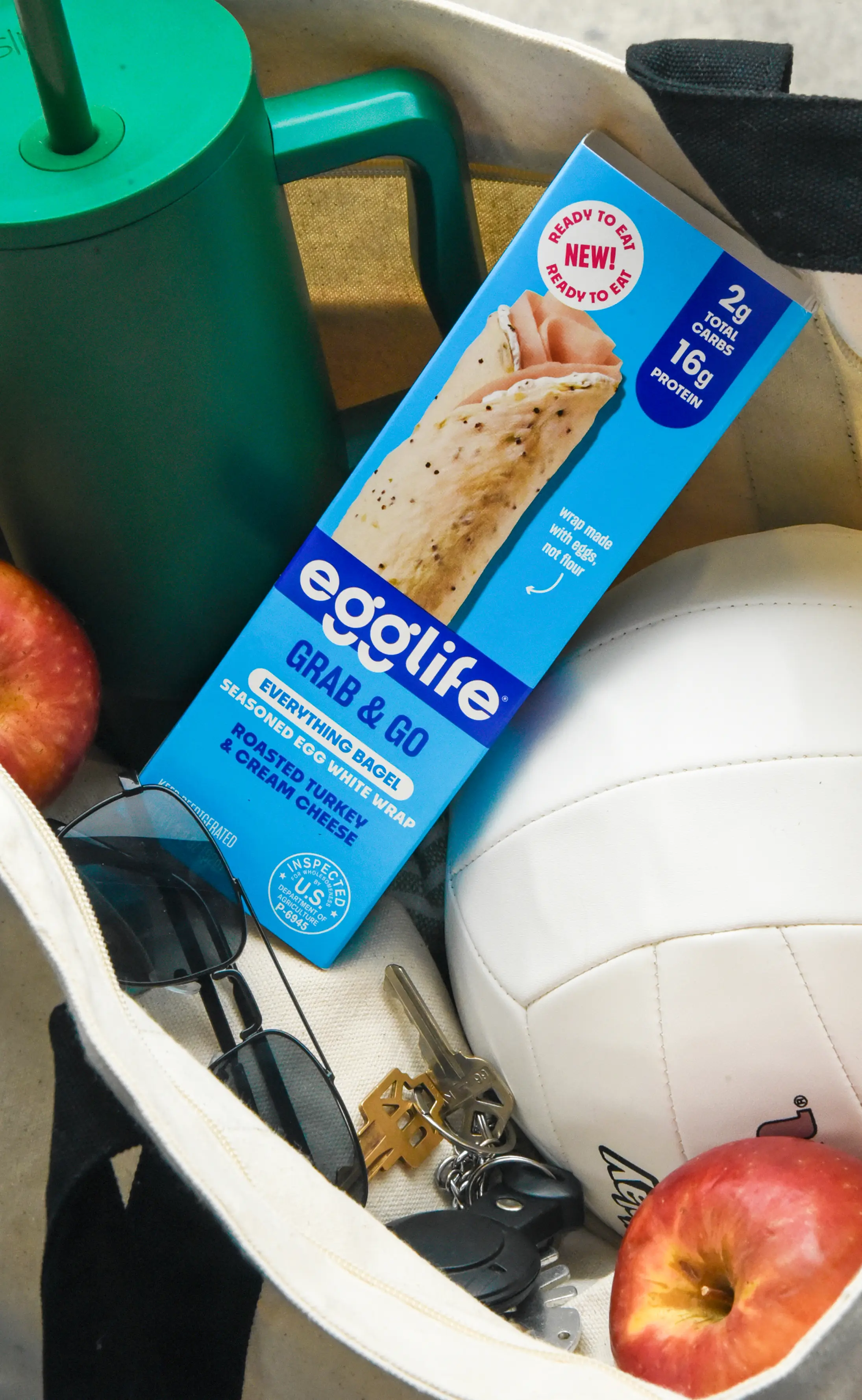 Canvas bag containing a white volleyball, a box of egglife Grab & Go bagel wrap, sunglasses, keys, and two red apples.