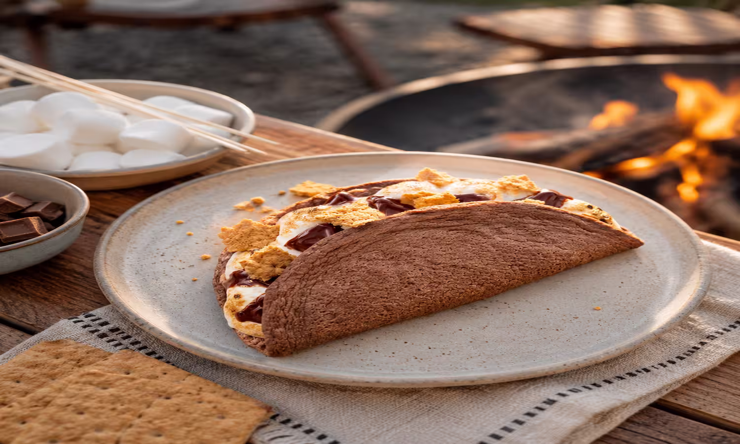 Chocolate egglife wrap folded into a s’mores with melted chocolate, toasted marshmallow, and graham cracker pieces, served by a campfire.