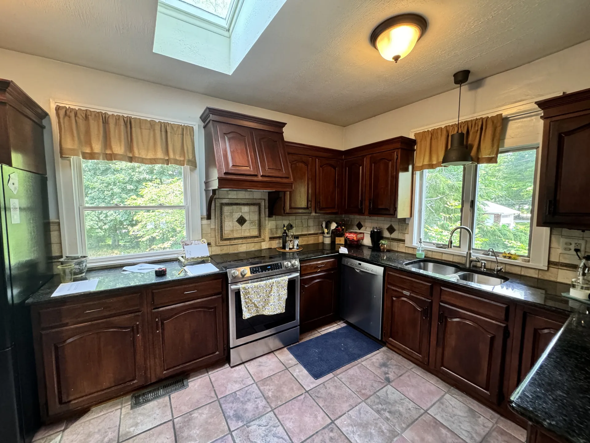 Before kitchen with dark cherry cabinets, tile floor, black countertops, and skylight with two windows.