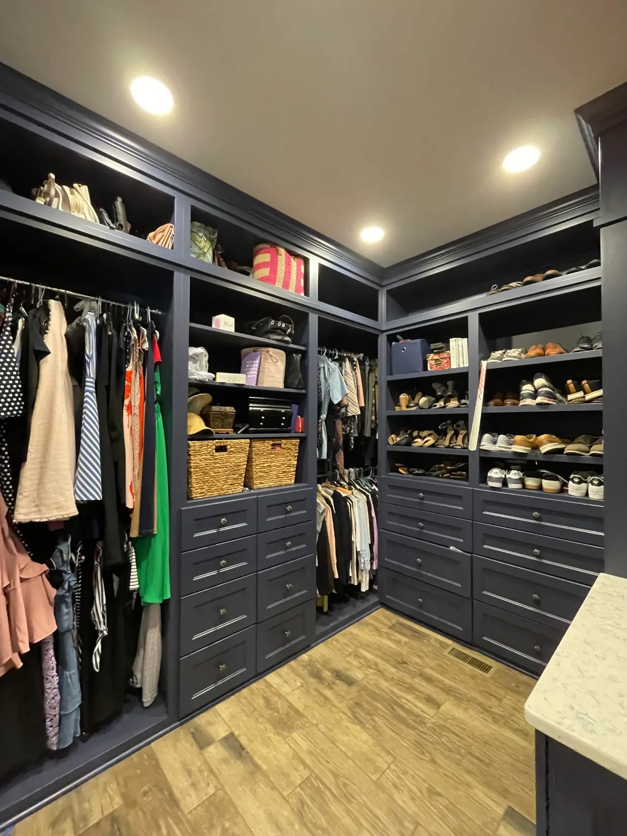 Walk-in closet with dark blue shelves and drawers, containing hanging clothes, woven baskets, handbags, hats, and multiple pairs of shoes on open shelves, illuminated by recessed ceiling lights and featuring wood-style flooring.