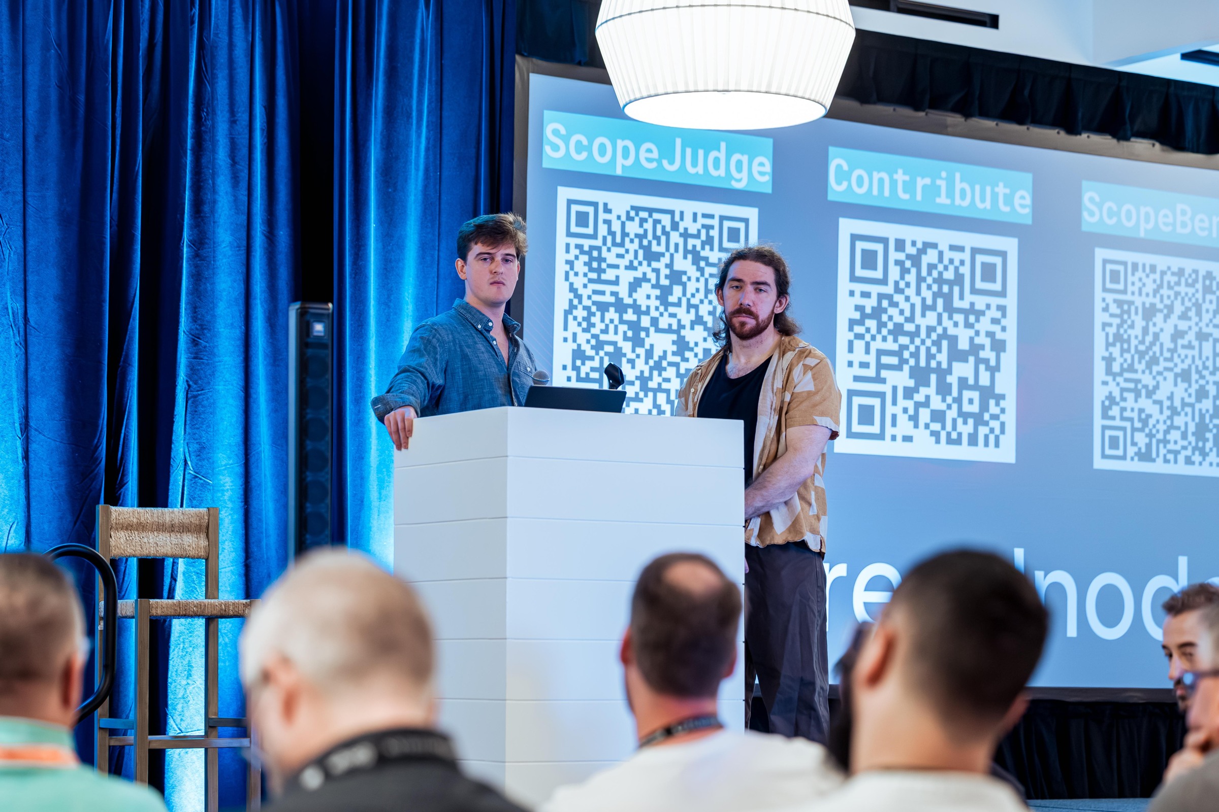 Two researchers presenting ScopeBench with contribution links projected behind.