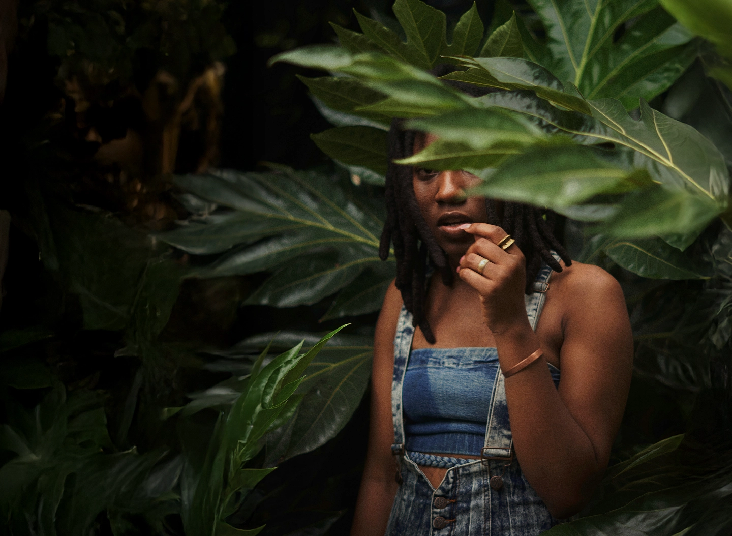 Young woman with dreadlocks partially hidden behind large green leaves, wearing denim overalls and jewelry.