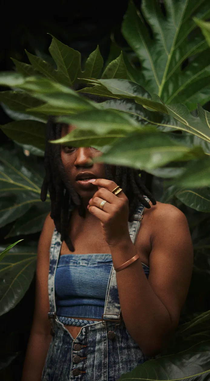 Woman with dreadlocks wearing denim overalls partially obscured by large green leaves.