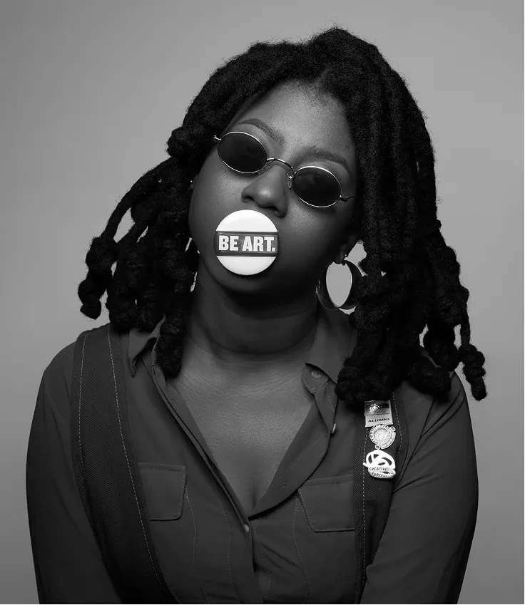 Black and white photo of a person with dreadlocks wearing round sunglasses and a button in their mouth that says 'BE ART.'