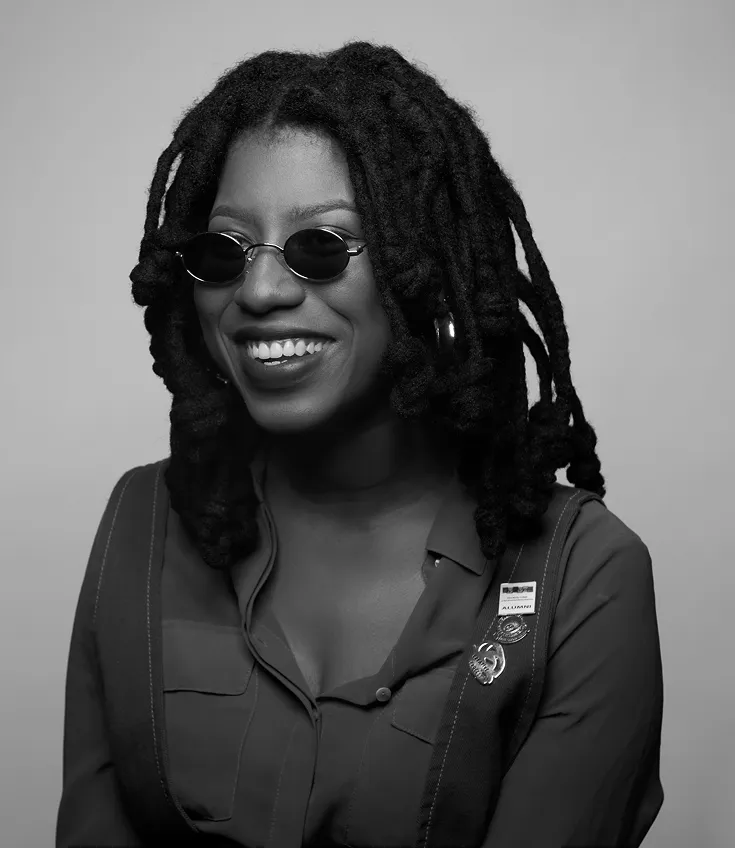 Black woman with dreadlocks wearing small round sunglasses and a buttoned shirt smiling against a plain background.