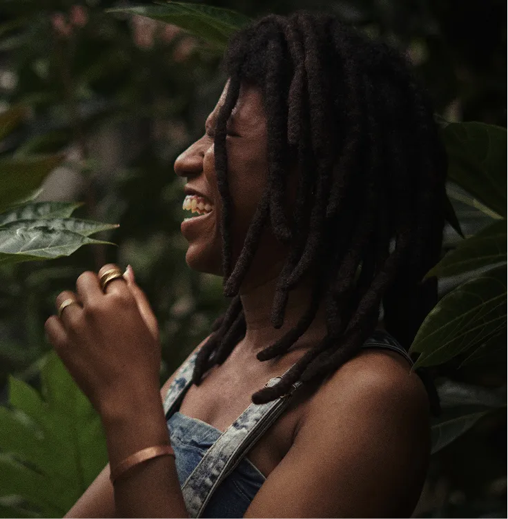 Profile of a woman with dreadlocks smiling and laughing amidst lush green foliage.