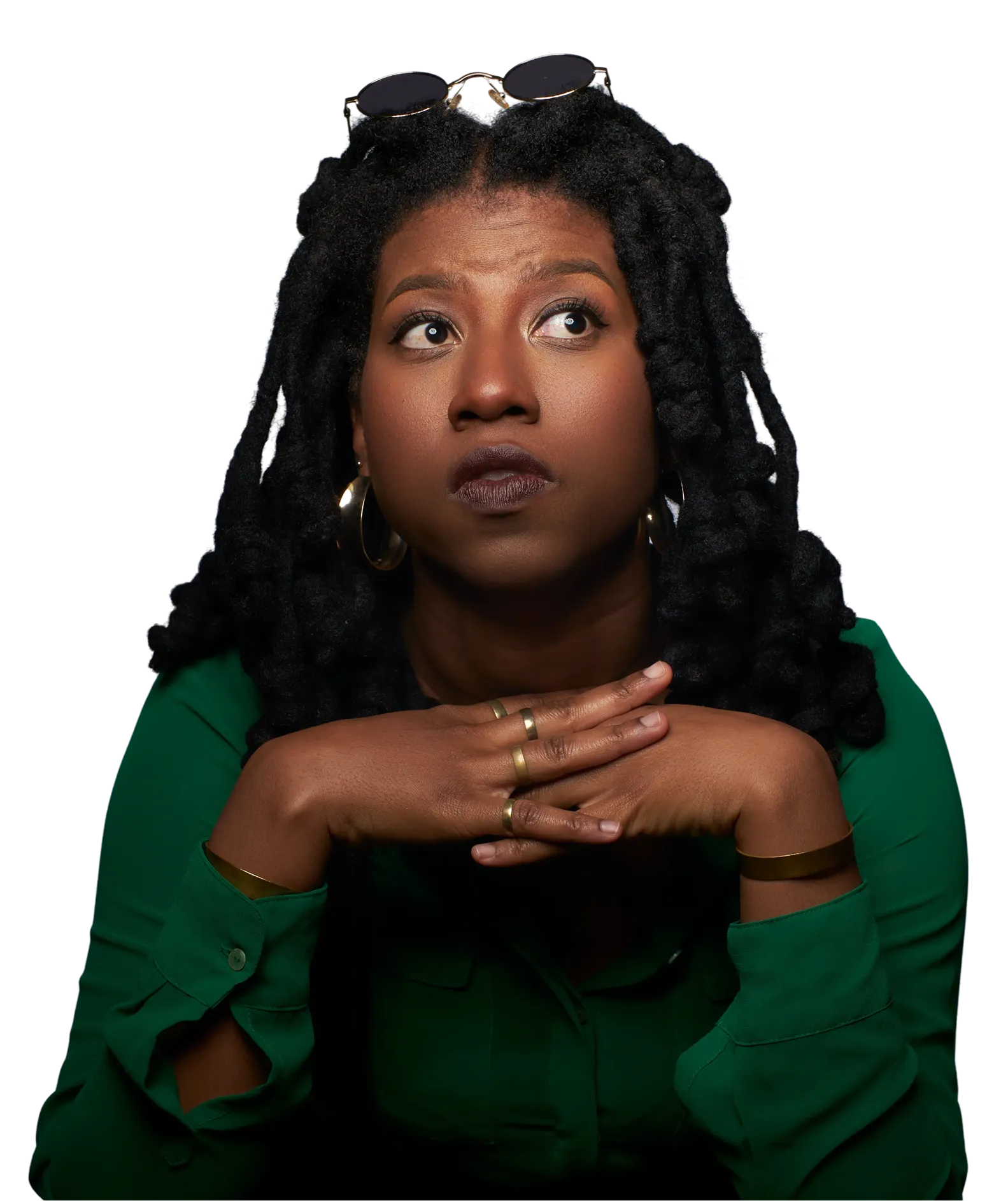 Thoughtful woman with dark curly hair and gold jewelry resting her chin on clasped hands wearing a green shirt and sunglasses on her head.