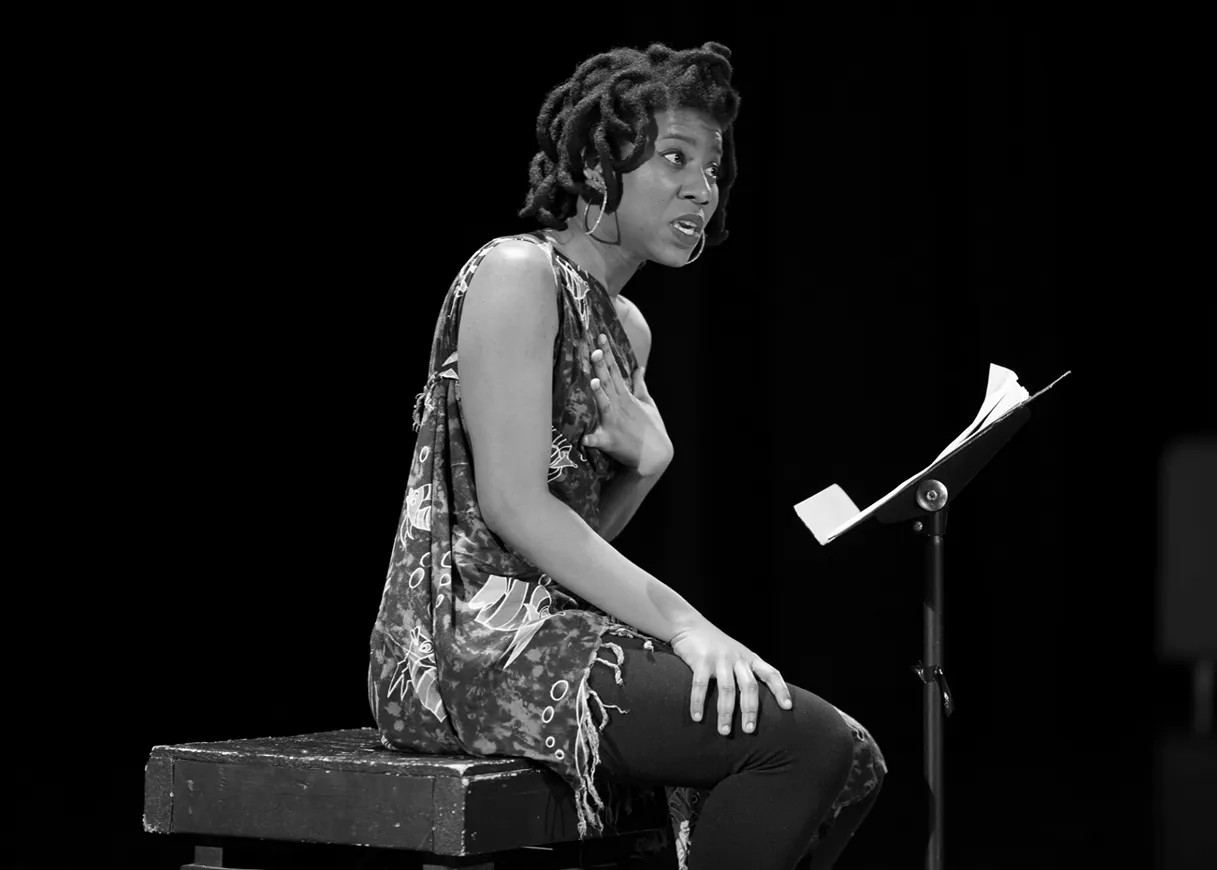 Black woman with styled hair and hoop earrings performing on stage, sitting on a bench beside a music stand with papers.