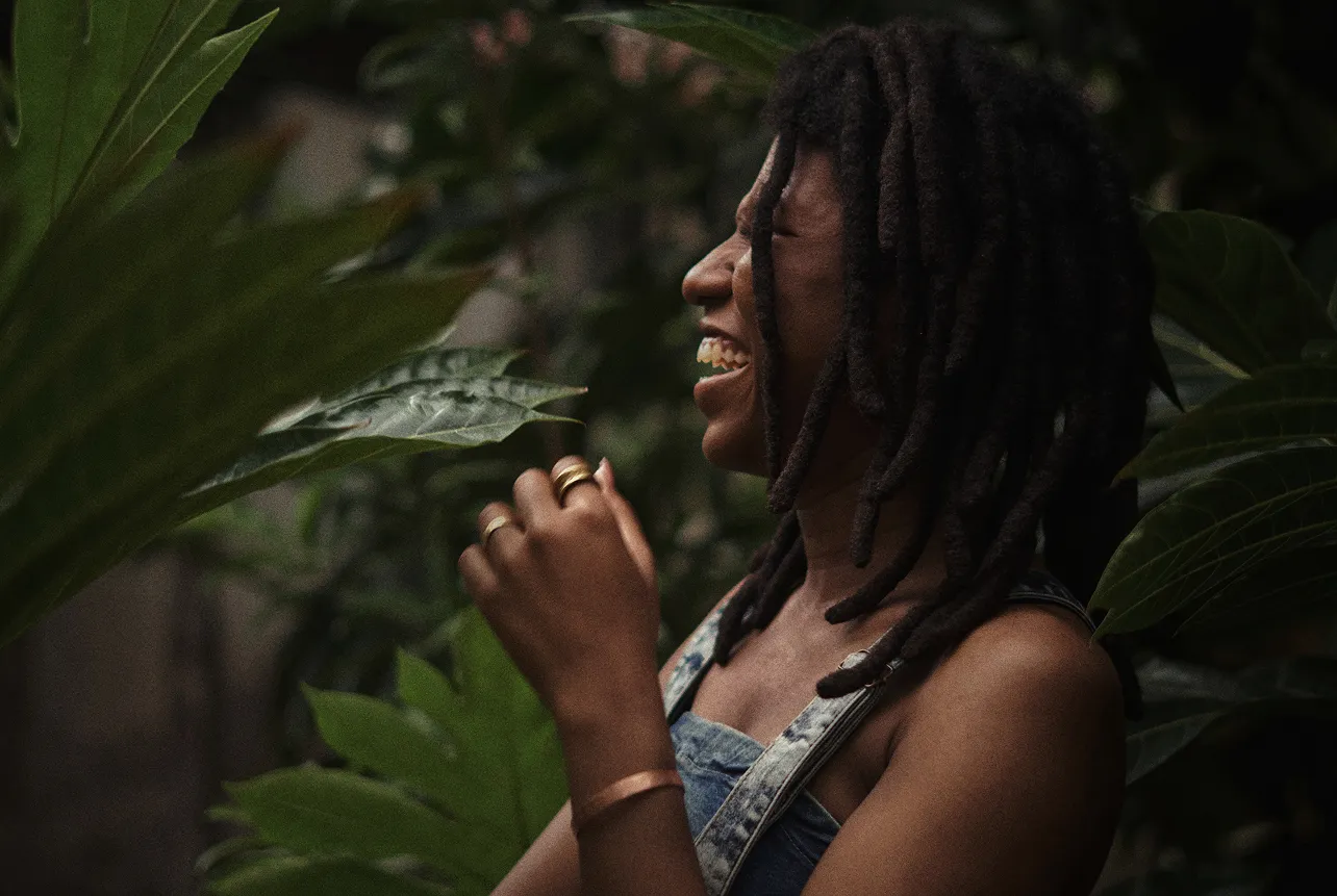 Woman with dreadlocks smiling and laughing among green leafy plants.