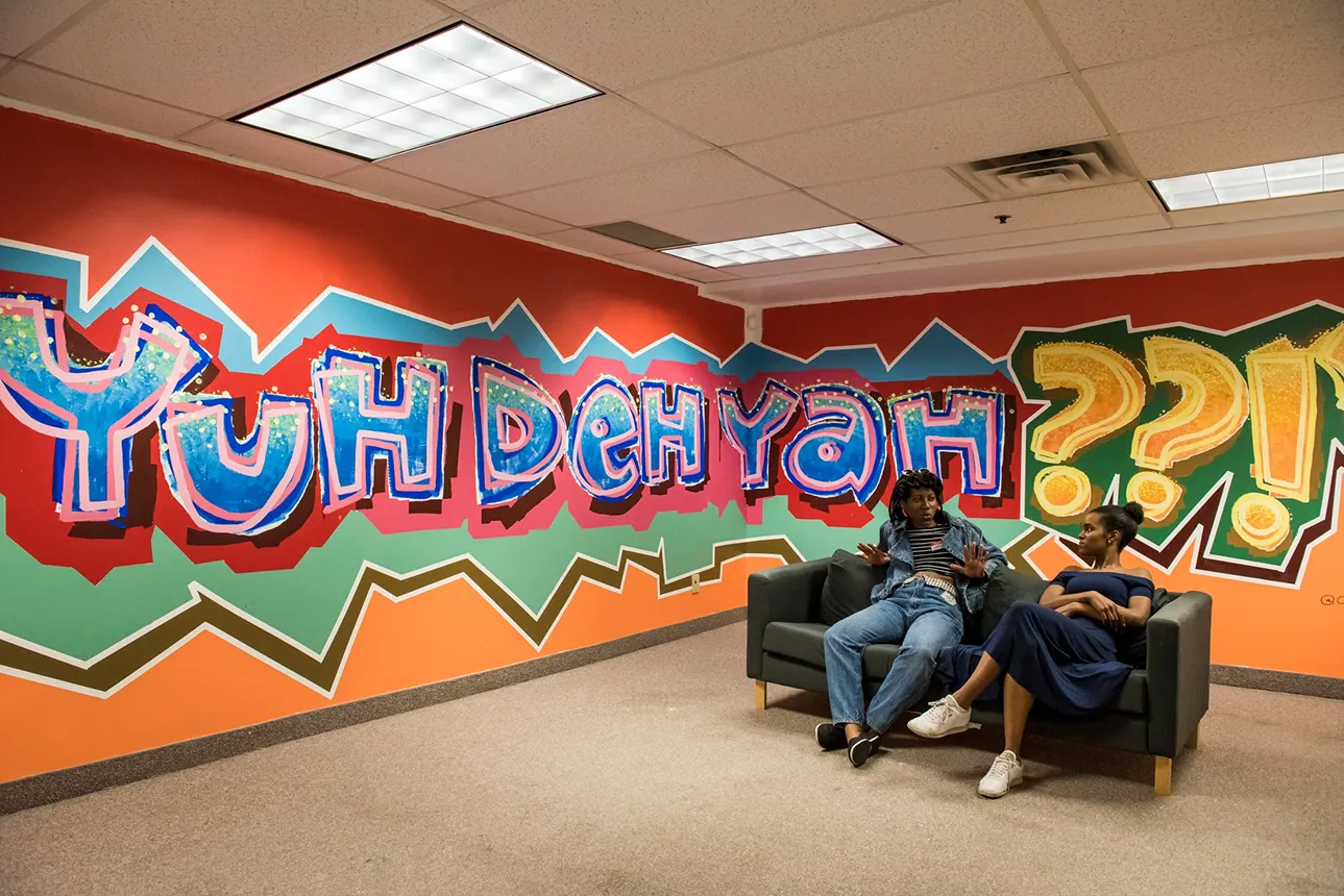 Two people sitting and talking on a couch in front of a colorful graffiti wall that says 'Yuh Deh Yah' with large question marks.