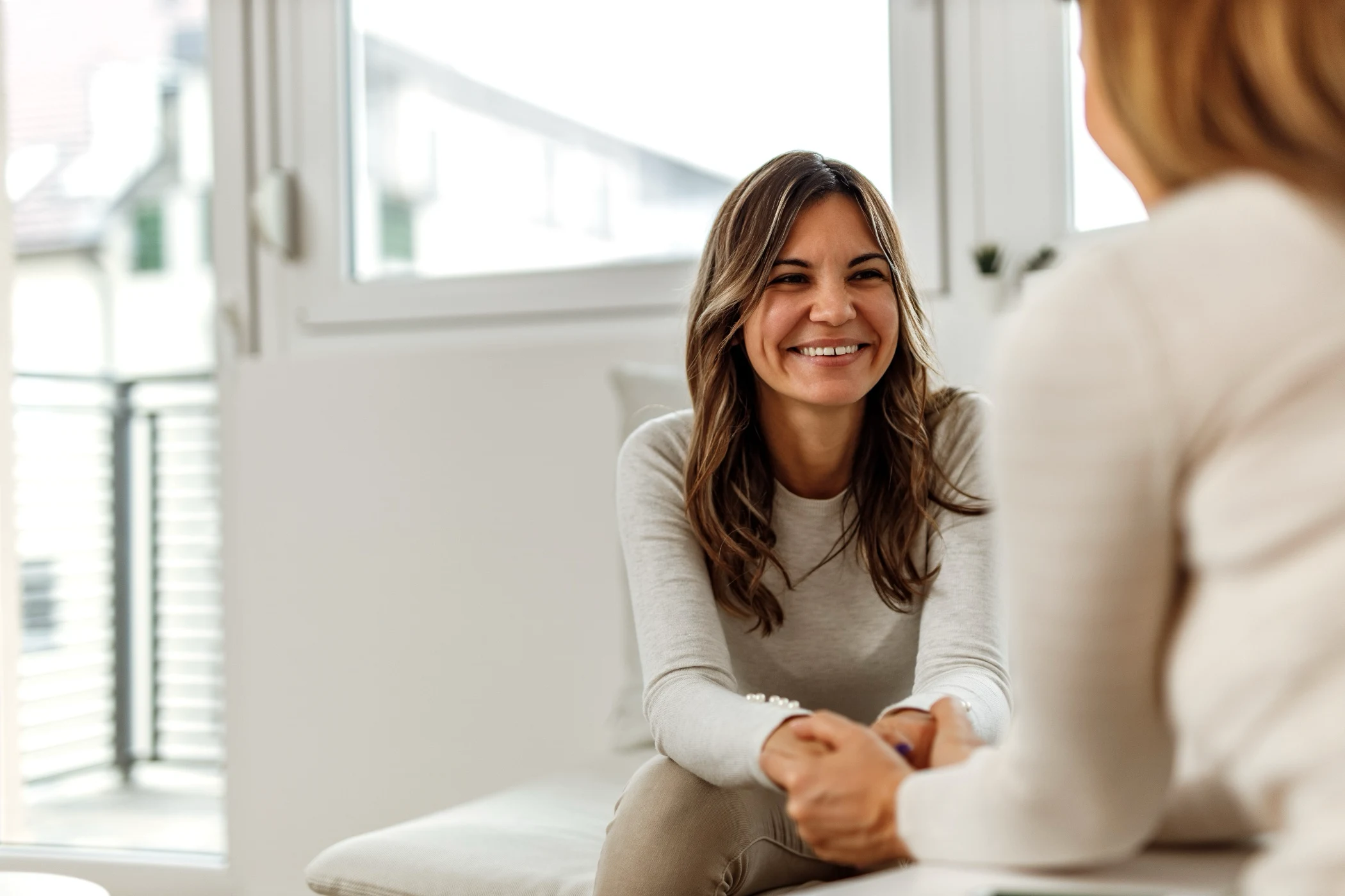 Smiling woman holding hands and talking with another person in a bright room.