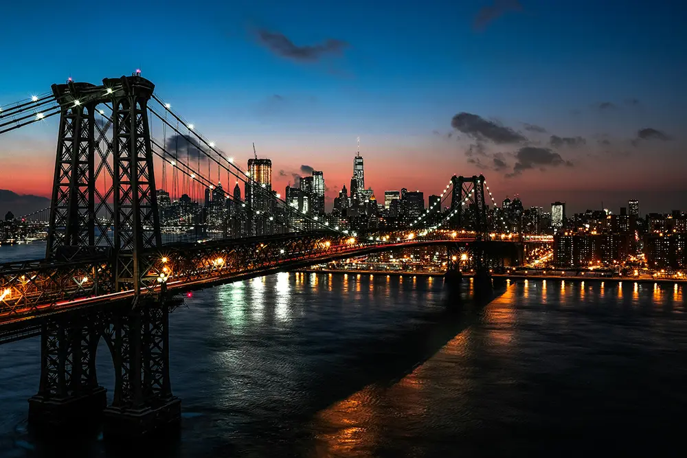 Pont suspendu illuminé traversant une rivière, devant la skyline de New York au crépuscule, ciel bleu orangé.