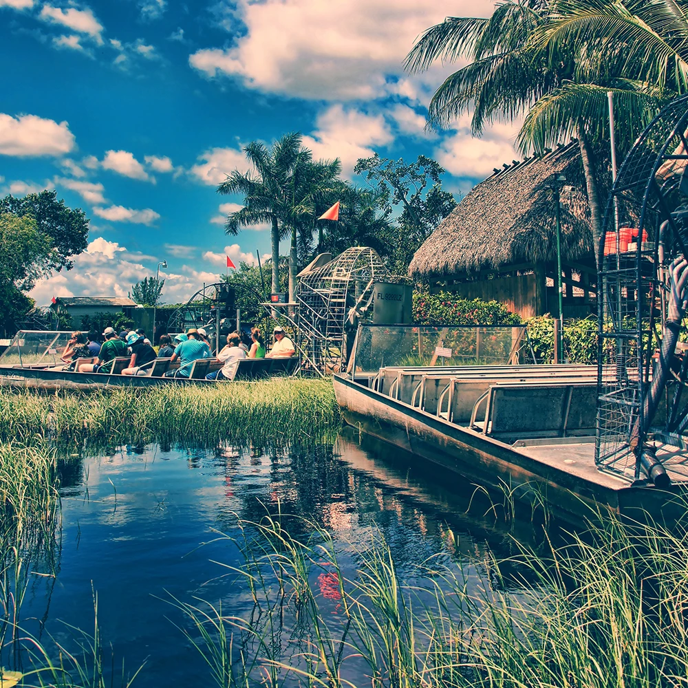 Touristes assis dans un hydroglisseur au bord d’un marais, près d’une hutte et de palmiers sous ciel bleu.