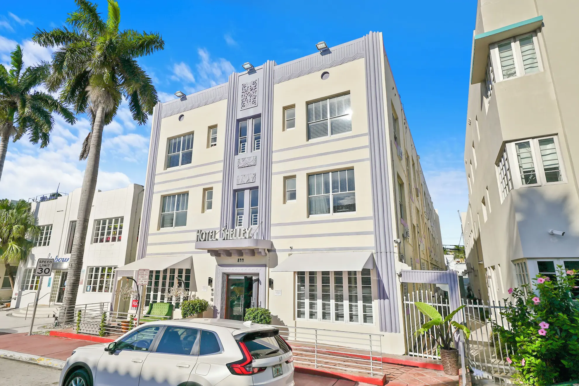 Façade art déco de l’hôtel Shelley sur rue ensoleillée, palmiers et SUV stationné au premier plan.
