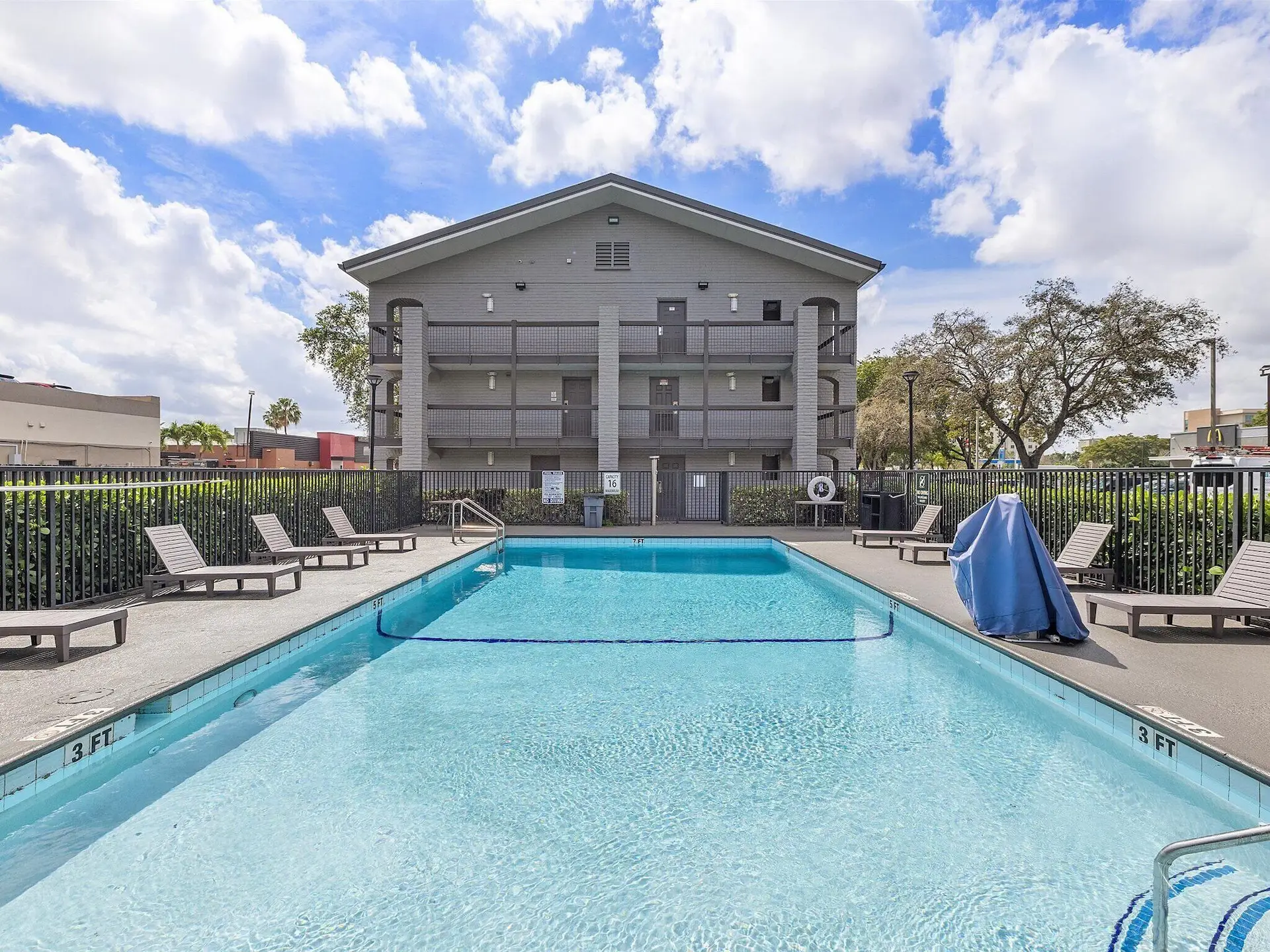 Piscine extérieure clôturée avec transats devant un immeuble gris, ciel partiellement nuageux.