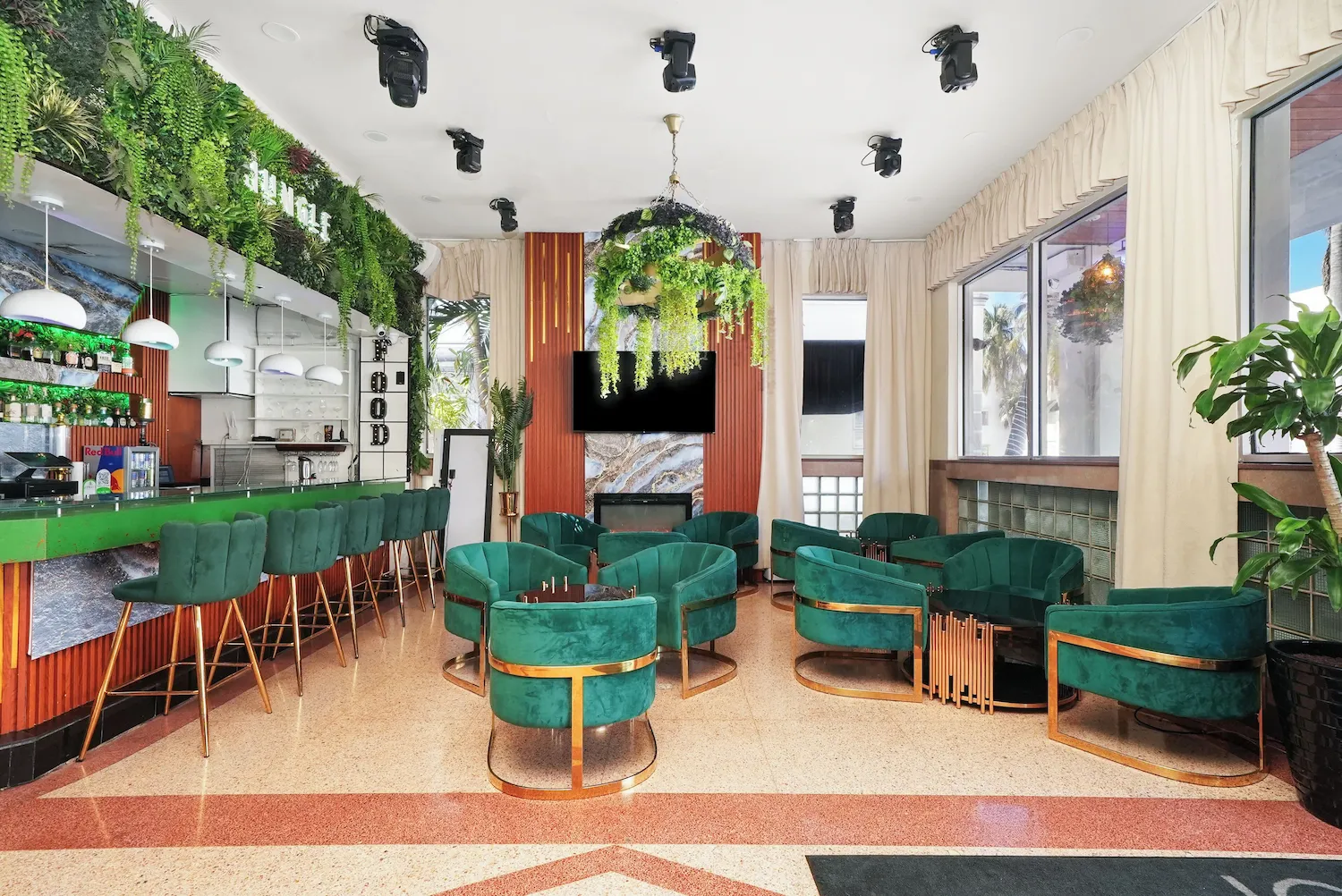 Salon de bar lumineux avec fauteuils vert émeraude et comptoir vert, plantes suspendues et grandes fenêtres.