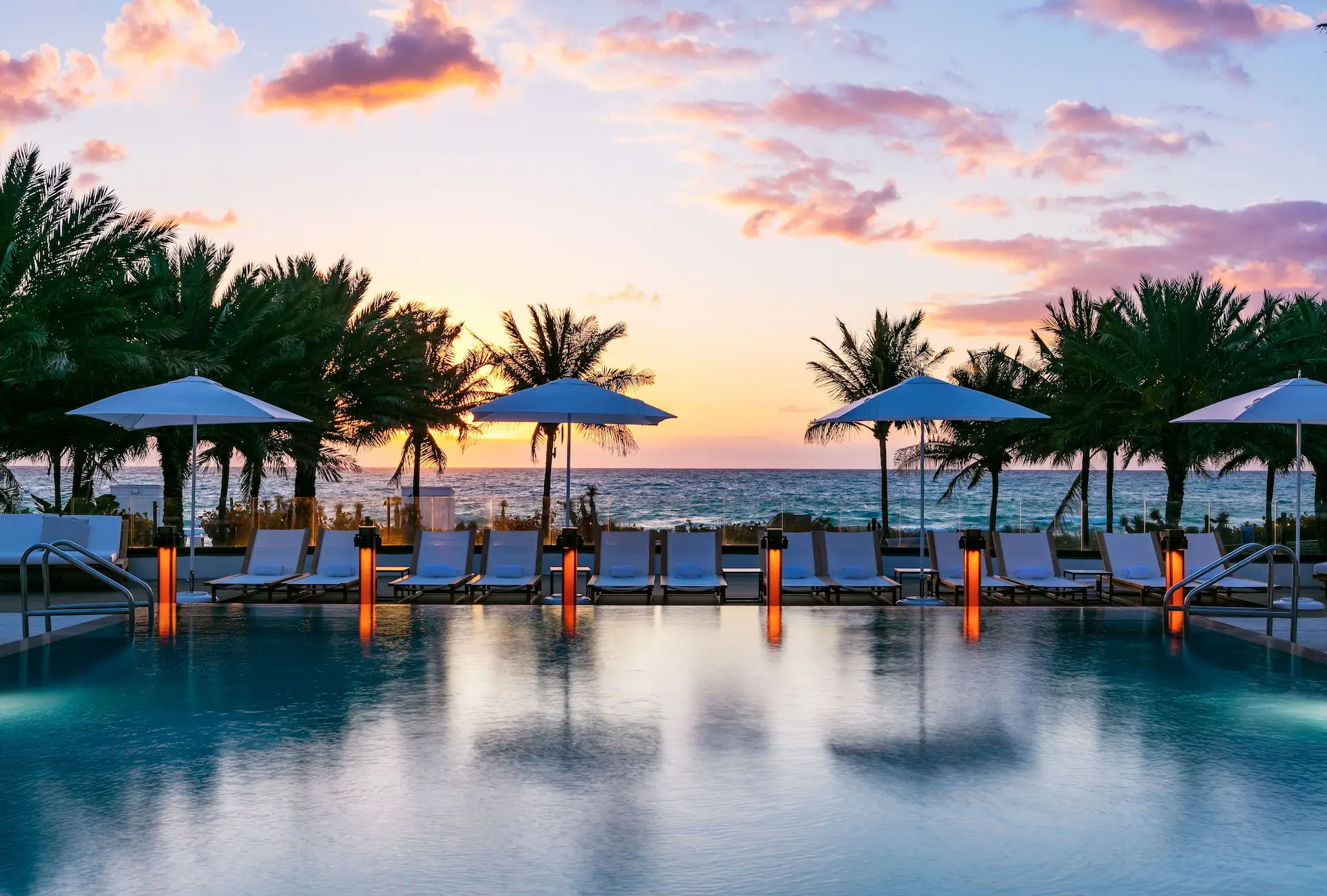 Piscine d’hôtel au bord de la mer au coucher du soleil, transats et parasols alignés sous des palmiers.