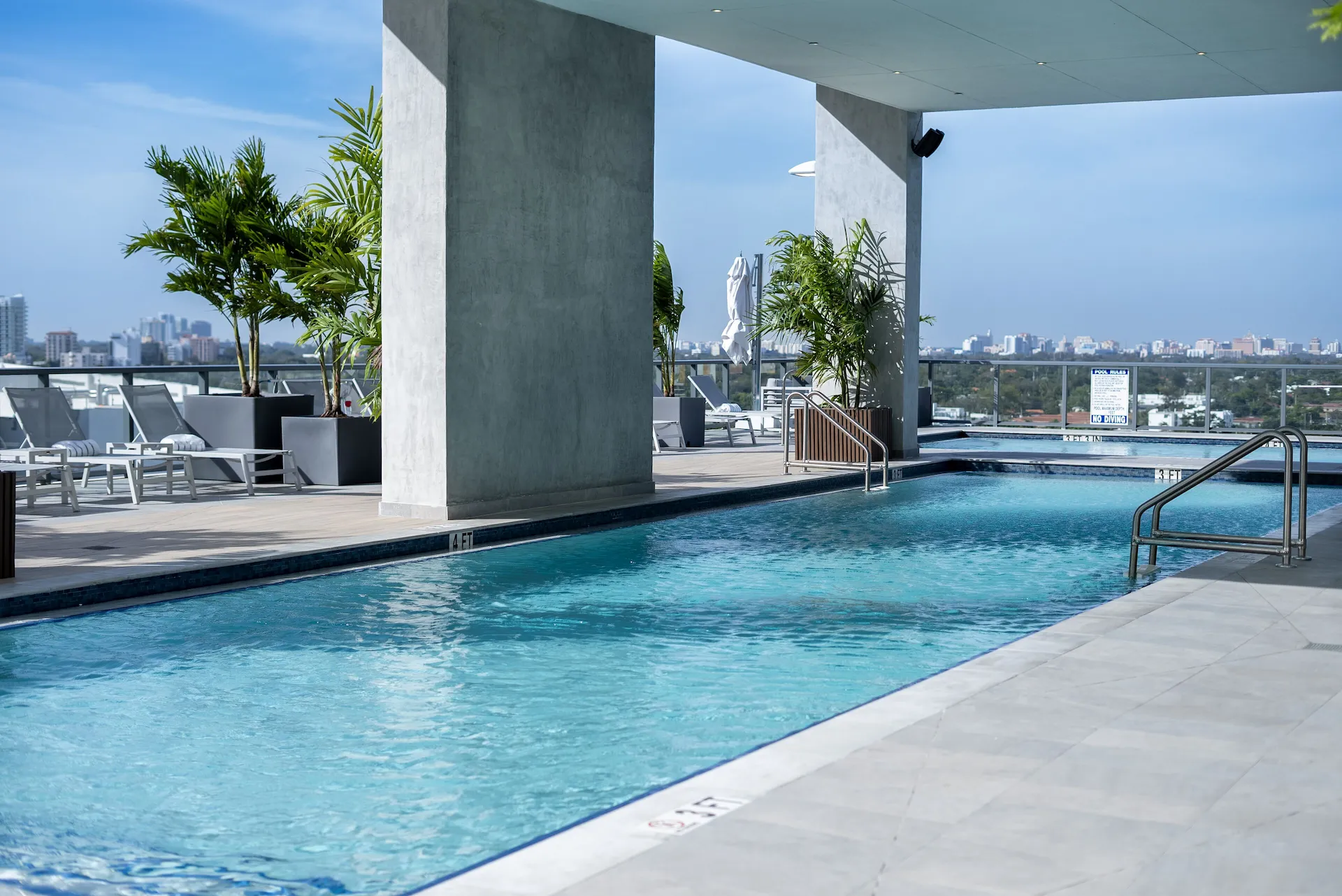 Piscine sur toit à demi couverte, colonnes en béton, palmiers en bacs et transats, vue urbaine lointaine par ciel bleu.