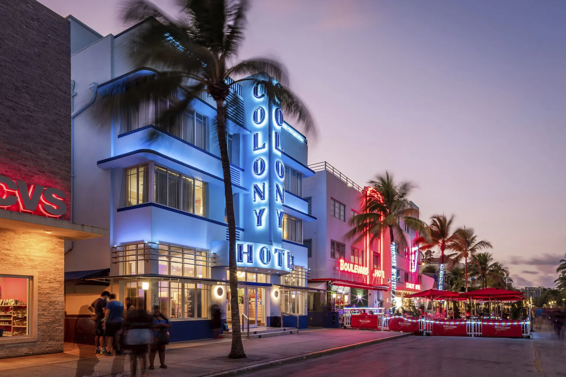 Façades art déco au néon du Colony Hotel et du Boulevard Hotel, palmiers et passants au crépuscule.