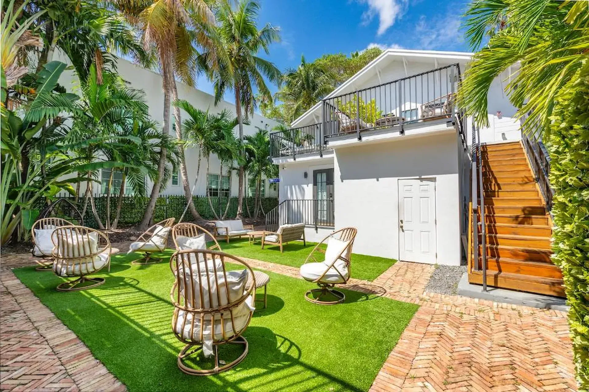 Cour avec palmiers, gazon synthétique, fauteuils en rotin, et maison blanche dotée d’une terrasse et d’un escalier bois.