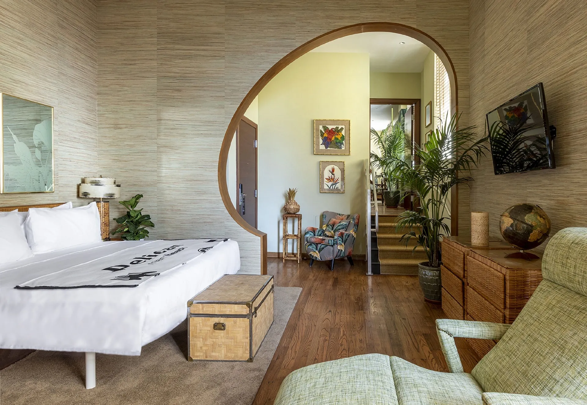 Chambre lumineuse avec lit blanc et coffre en rotin, grande arche ovale, plantes, fauteuil fleuri et TV murale.