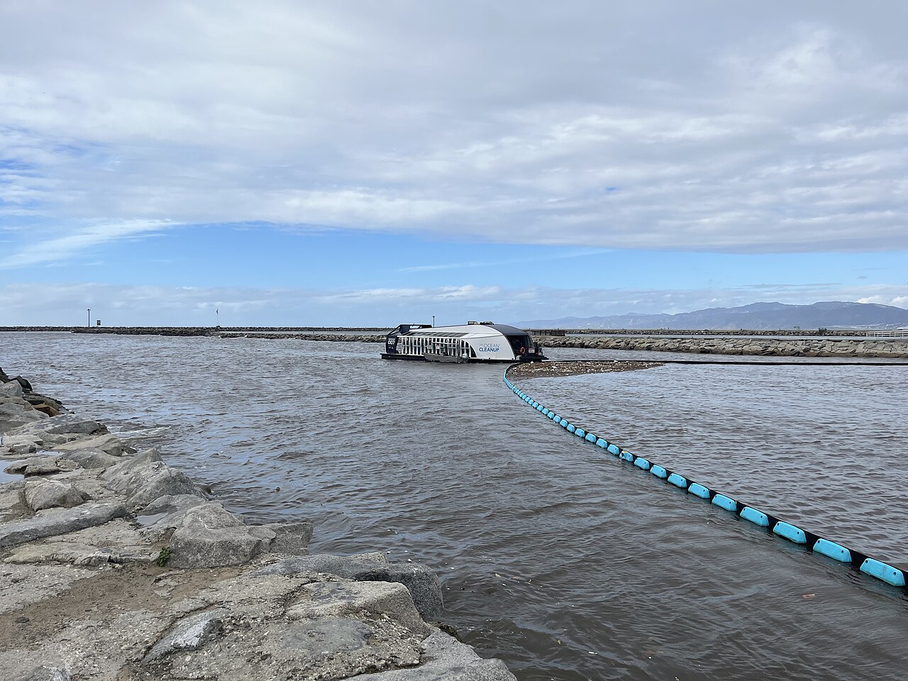 The Ocean Cleanup's Interceptor vessel at a river mouth with a floating boom line collecting debris