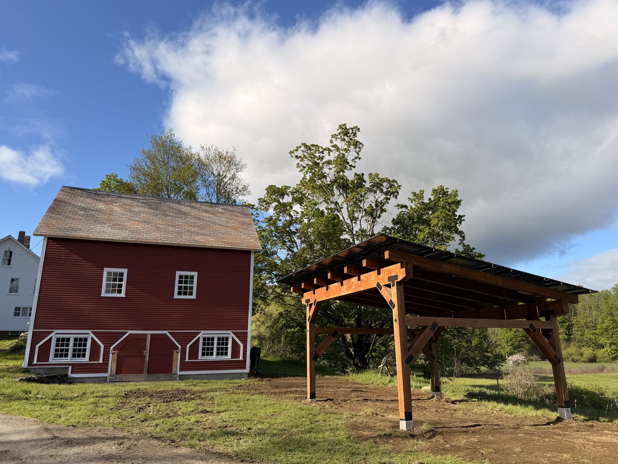 A Kingdom Sun timber-frame solar carport beside a classic red Vermont barn in Dummerston, panels overhead, blue sky