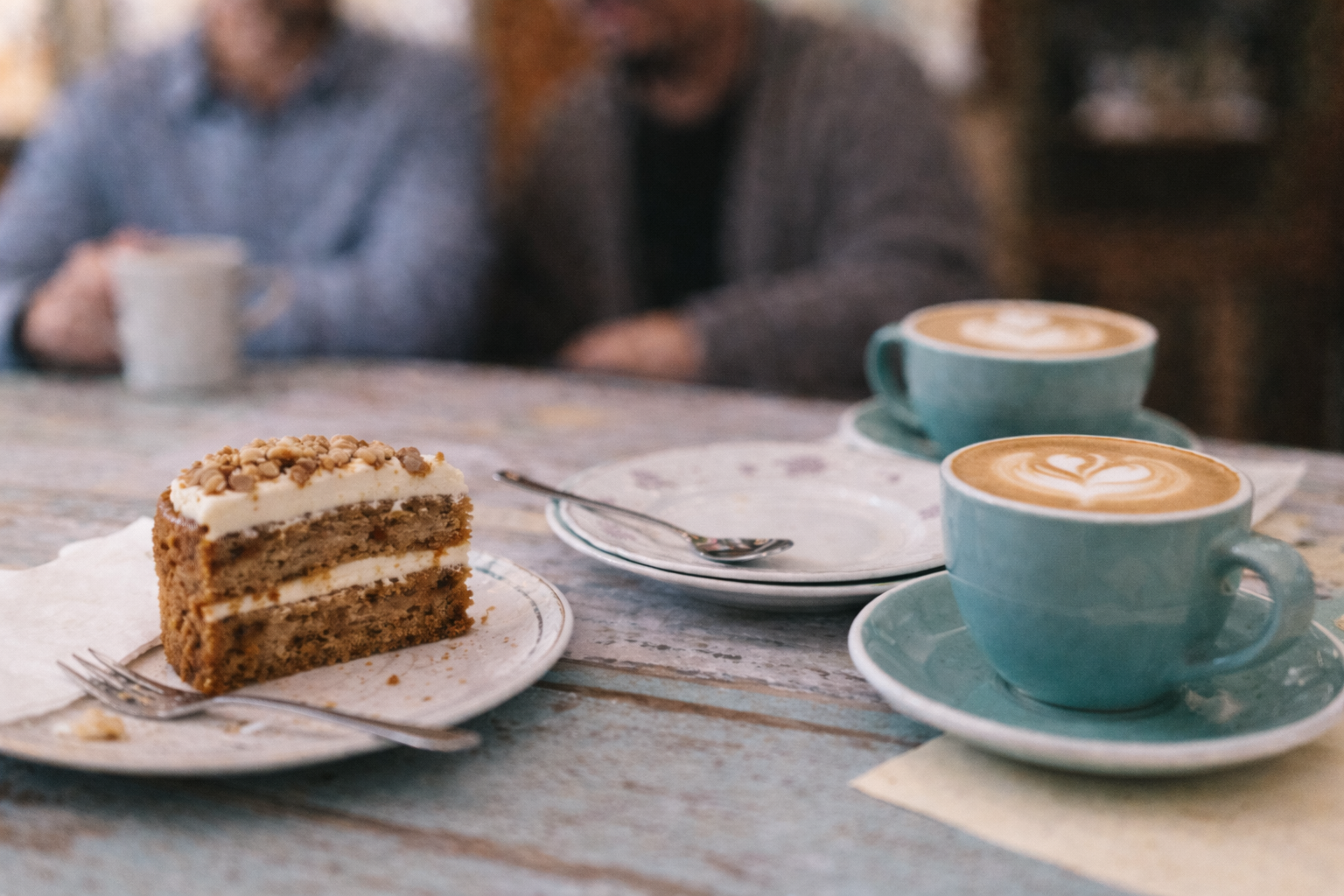 Two lattes and a slice of layered carrot cake on a rustic table.