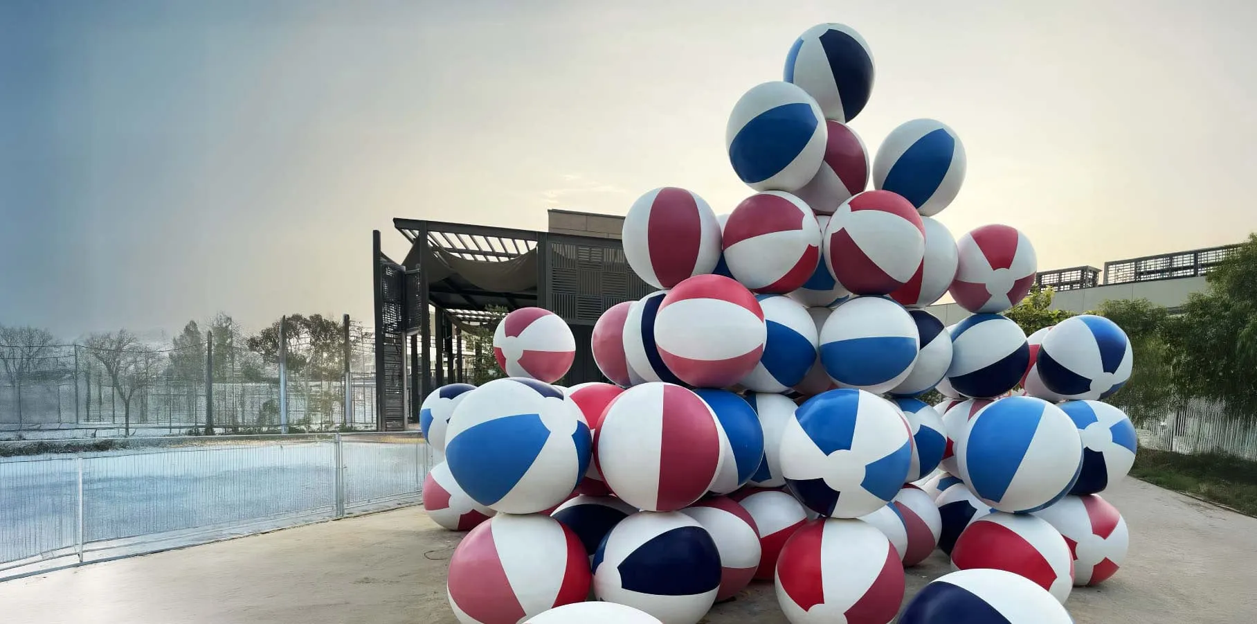Large pyramid stack of red, white, and blue beach balls outdoors near a pool and fence at sunset.