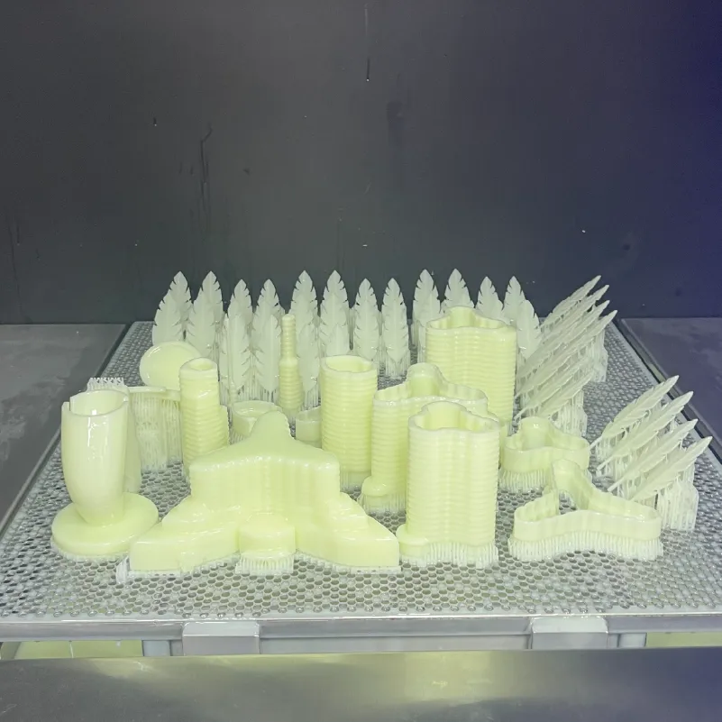 Multiple pale yellow 3D printed objects including trees and abstract shapes on a metal grid platform.