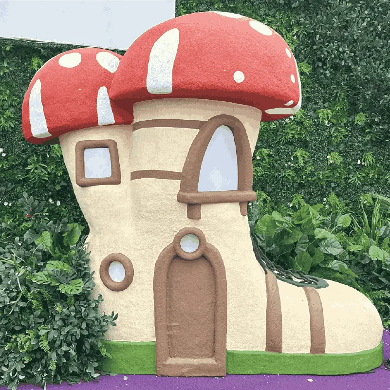 Sculpture of a whimsical boot-shaped house with red mushroom caps and greenery around.