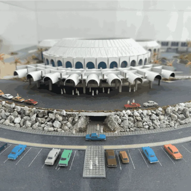Scale model of a round stadium with cars and small human figures in front, surrounded by a rocky wall.